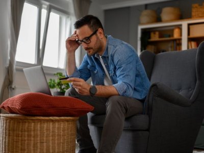 Man sitting at laptop with credit card in hand looking worried. Adobe.